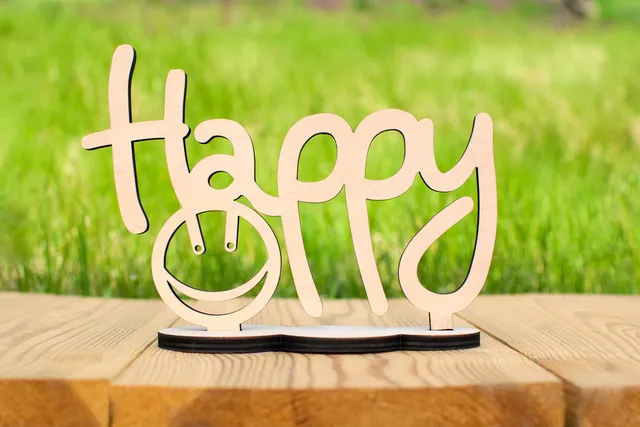 Cuvântul „Happy” decupat în lemn, pe o masă, în fața unui câmp verde
